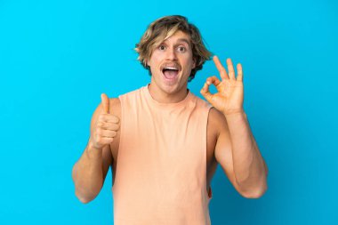 Yakışıklı sarışın adam mavi arka planda izole edilmiş tamam işareti ve baş parmak işareti gösteriyor