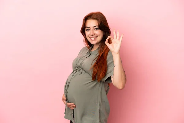 若いですRedhead Caucasian女性隔離上のピンクの背景妊娠とやってOkサイン — ストック写真