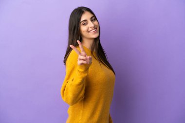 Mor arka planda izole edilmiş genç beyaz kadın gülümsüyor ve zafer işareti gösteriyor.