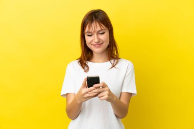 Kızıl saçlı kız sarı arkaplanda izole edilmiş. Cep telefonuyla mesaj ya da e-posta gönderiyor.