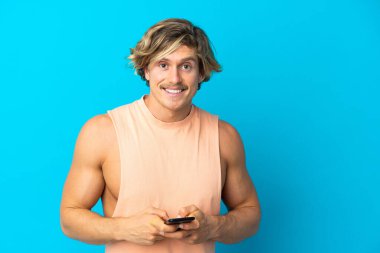 Yakışıklı sarışın adam mavi arkaplanda izole edilmiş. Cep telefonuyla mesaj gönderiyor.