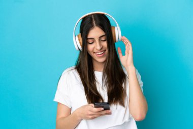 Genç beyaz kadın mavi arka planda izole edilmiş müzik dinliyor ve cep telefonu arıyor.