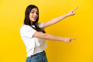 Sarı arka planda izole edilmiş genç, beyaz bir kadın parmak göstererek bir ürün sunuyor.