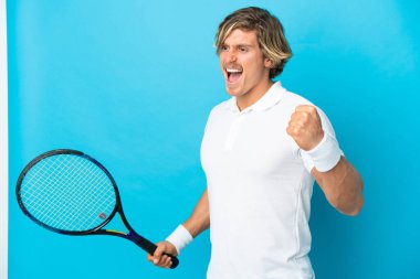 Genç sarışın adam mavi arka planda izole edilmiş tenis oynuyor ve zaferi kutluyor.