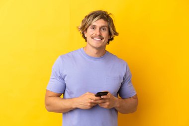Sarışın yakışıklı adam sarı arka planda izole edilmiş cep telefonuyla mesaj gönderiyor.