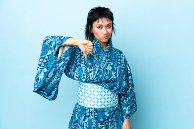 İzole edilmiş mavi arka planda kimono giyen genç bir kadın olumsuz ifadeyle baş parmağını aşağı gösteriyor.