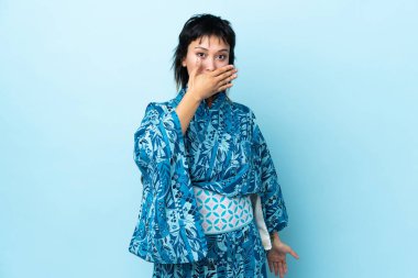 İzole edilmiş mavi arka planda kimono giyen genç bir kadın ağızlarını eliyle kapatıyor.
