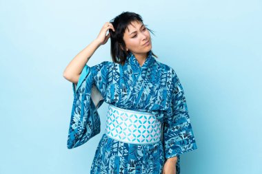 İzole edilmiş mavi arka planda kimono giyen genç bir kadın kafasını kaşırken şüpheye düşüyor.
