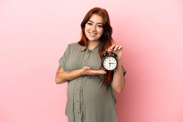 Młoda Ruda Biały Kobieta Odizolowany Różowy Tle Ciąży Trzymając Zegar — Zdjęcie stockowe
