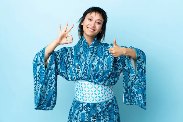 若いです女性の上に着物を着て隔離された青の背景を示すOkサインと親指アップジェスチャー — ストック写真
