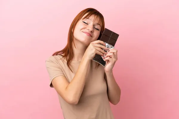 Roodharige Meisje Geïsoleerd Roze Achtergrond Nemen Van Een Chocolade Tablet — Stockfoto