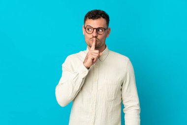 Mavi arka planda izole edilmiş beyaz, yakışıklı genç bir adam sessiz bir jest yapıyor. Parmağını ağzına sokuyor.