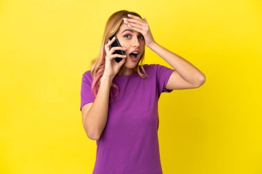 İzole edilmiş sarı arka plan üzerinde cep telefonu kullanan genç bir kadın yan tarafa bakarken sürpriz jest yapıyor.