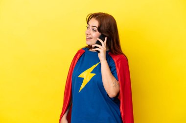 Süper kahraman kızıl saçlı kadın sarı arka planda izole edilmiş cep telefonuyla biriyle sohbet ediyor.