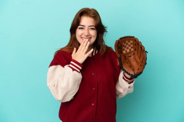 Beyzbol oynayan genç kızıl saçlı kadın mavi arka planda izole edilmiş mutlu ve ağzı kapalı gülümsüyor.