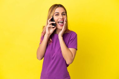 İzole edilmiş sarı arka planda cep telefonu kullanan genç bir kadın ağzı açık bağırıyor.