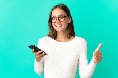 İzole edilmiş mavi arka plan üzerinde genç bir kadın başparmak kaldırırken cep telefonu kullanıyor.