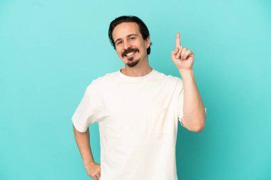 Kafkasyalı genç adam mavi arka planda izole edildi ve en iyinin işareti olarak parmağını kaldırdı.