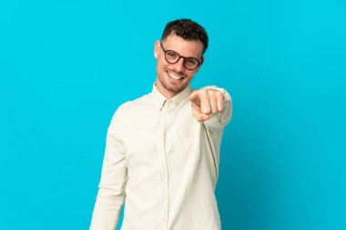 Genç beyaz yakışıklı adam mavi arka planda izole edilmiş mutlu bir ifadeyle ön tarafı işaret ediyor.