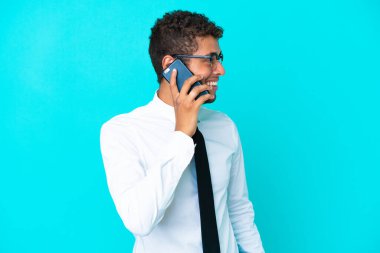 Genç Brezilyalı iş adamı mavi arka planda izole edilmiş cep telefonuyla biriyle sohbet ediyor.