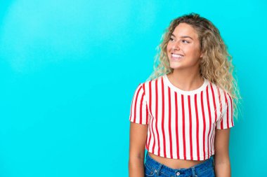 Kıvırcık saçlı kız mavi arka planda izole edilmiş, yan tarafa bakıyor ve gülümsüyor.