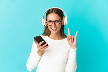 İzole edilmiş mavi arka planda gezici bir rock hareketi yapan genç bir kadın müzik dinliyor.
