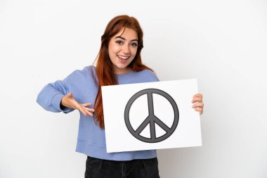 Genç kızıl saçlı kadın beyaz arka planda izole edilmiş. Elinde barış sembolü olan bir pankart var ve onu işaret ediyor.