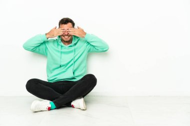 Genç, yakışıklı, beyaz bir adam yerde oturuyor. Gözlerini elleriyle kapatıyor.