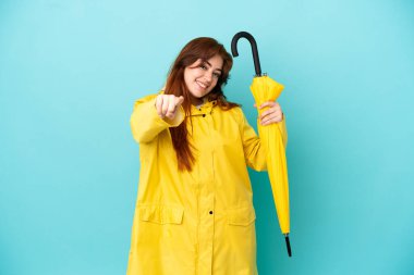 Mavi arka planda şemsiyesi olan kızıl saçlı kadın mutlu bir ifadeyle ön tarafı işaret ediyor.