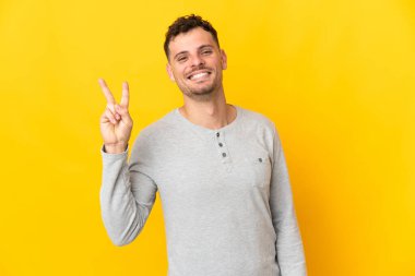 Sarı arka planda izole edilmiş yakışıklı genç adam gülümsüyor ve zafer işareti gösteriyor.