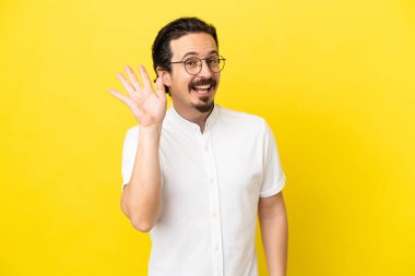 Sarı arka planda izole edilmiş genç beyaz adam mutlu bir ifadeyle selamlıyor.