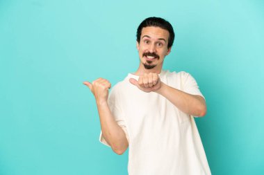 Mavi arka planda izole edilmiş genç beyaz adam bir ürünü sunmak için kenarı işaret ediyor.