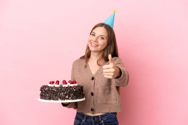 Elinde doğum günü pastası tutan Litvanyalı genç bir kadın pembe arka planda izole edilmiş, başparmakları havada çünkü iyi bir şey oldu.
