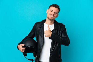 Motosiklet kasklı beyaz bir adam mavi arka planda izole edilmiş bir şekilde baş parmak işareti yapıyor.