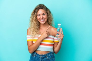 Mavi arka planda izole edilmiş su şişesi olan genç sarışın kadın bir ürünü sunmak için yan tarafı işaret ediyor.