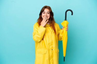 Mavi arka planda şemsiyesi olan kızıl saçlı kadın mutlu ve gülümsüyor.