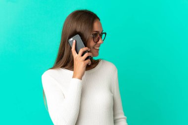 İzole edilmiş mavi arka planı olan genç bir kadın cep telefonuyla sohbet ediyor.