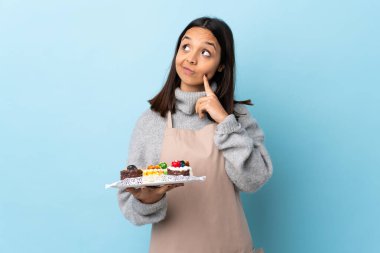 Pasta şefi, mavi arka planda büyük bir pasta tutuyor. Yukarı bakarken bir fikir düşünüyor..