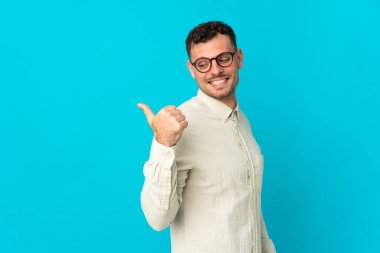 Mavi arka planda izole edilmiş beyaz, yakışıklı genç bir adam bir ürün sunmak için kenarı işaret ediyor.