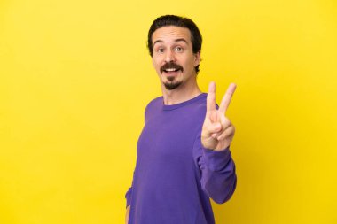 Sarı arka planda izole edilmiş genç beyaz adam gülümsüyor ve zafer işareti gösteriyor.