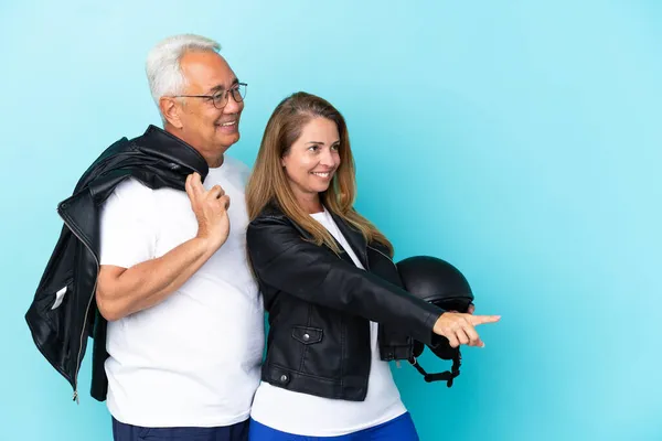 Middle age bikers couple with a motorcycle helmet isolated on blue background pointing to the side to present a product