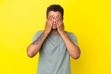 Genç ve yakışıklı Brezilyalı adam sarı arka planda izole edilmiş, gözleri elle kaplanmış.