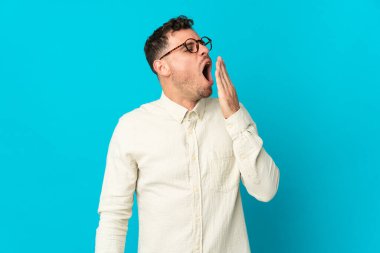 Beyaz tenli yakışıklı adam mavi arka planda izole edilmiş esniyor ve ağzı açık bir şekilde elleriyle ağzını kapatıyor.