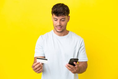 Genç beyaz yakışıklı adam sarı arka planda izole edilmiş cep telefonuyla kredi kartıyla alışveriş yapıyor.