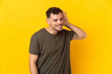 Beyaz tenli yakışıklı adam sarı arka planda izole edilmiş bir şekilde elini kulağına koyarak bir şeyler dinliyor.