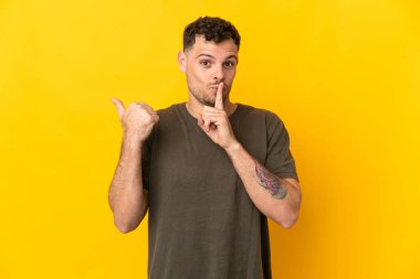Sarı arka planda izole edilmiş genç, beyaz, yakışıklı bir adam. Yan tarafı işaret ediyor ve sessizlik hareketi yapıyor.
