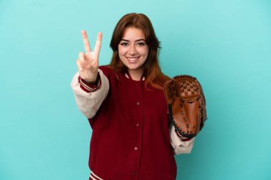 Beyzbol oynayan genç kızıl saçlı kadın mavi arka planda izole edilmiş gülümsüyor ve zafer işareti gösteriyor.