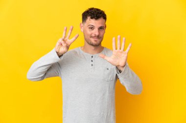 Sarı arka planda izole edilmiş beyaz, yakışıklı genç bir adam sekiz parmaklı sayıyor.