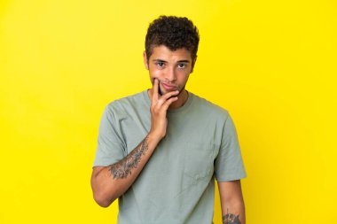 Genç yakışıklı Brezilyalı adam sarı arka planda izole edilmiş ve düşünüyor.