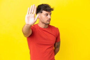 Sarı arka planda izole edilmiş beyaz, yakışıklı genç adam dur hareketi yapıyor ve hayal kırıklığına uğruyor.